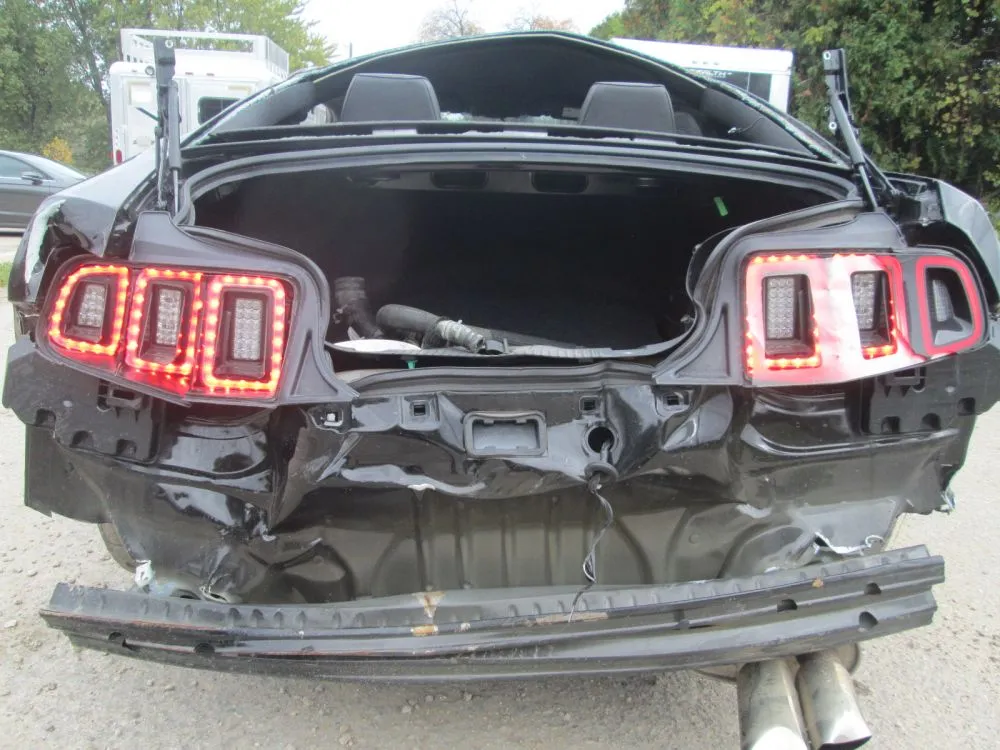 A black Mustang with severe rear-end collision damage in the Fine's Body Shop lot, awaiting repair